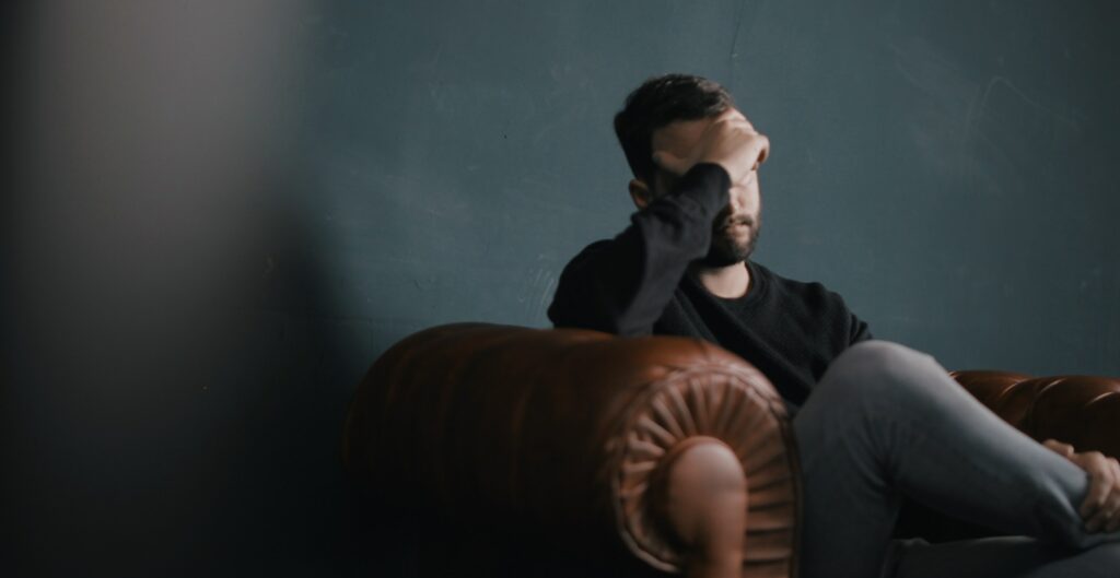 A man sits on a couch with his hand on his face to represent symptoms of heroin withdrawal.