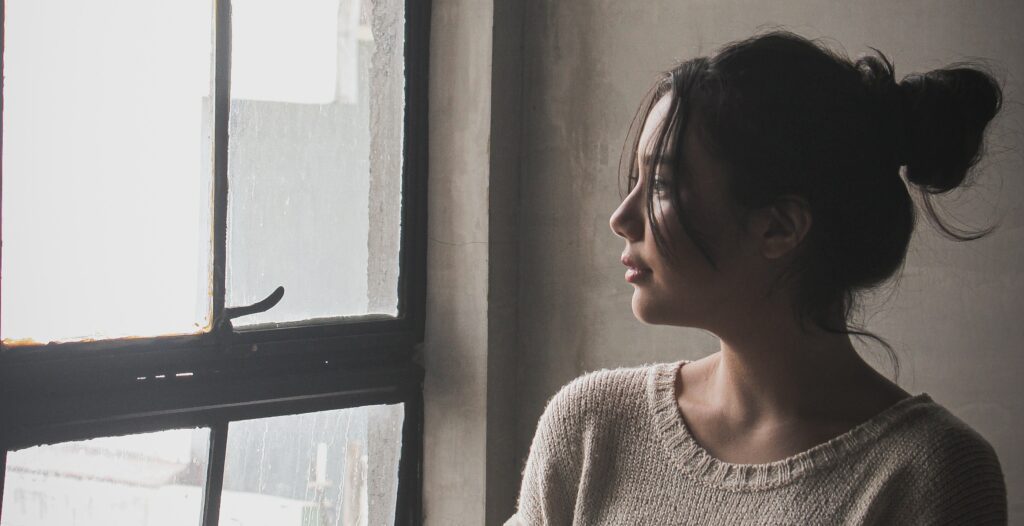 a woman looks out a window to represent the question, "how long does alcohol withdrawal last?".